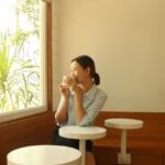 A woman sits alone by a sunny cafe window, smiling as she sips her coffee