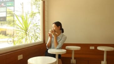 A woman sits alone by a sunny cafe window, smiling as she sips her coffee