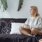 A middle-aged woman sits cross-legged on a sofa holding a cup of coffee, looking thoughtfully out of frame