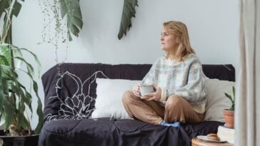 A middle-aged woman sits cross-legged on a sofa holding a cup of coffee, looking thoughtfully out of frame